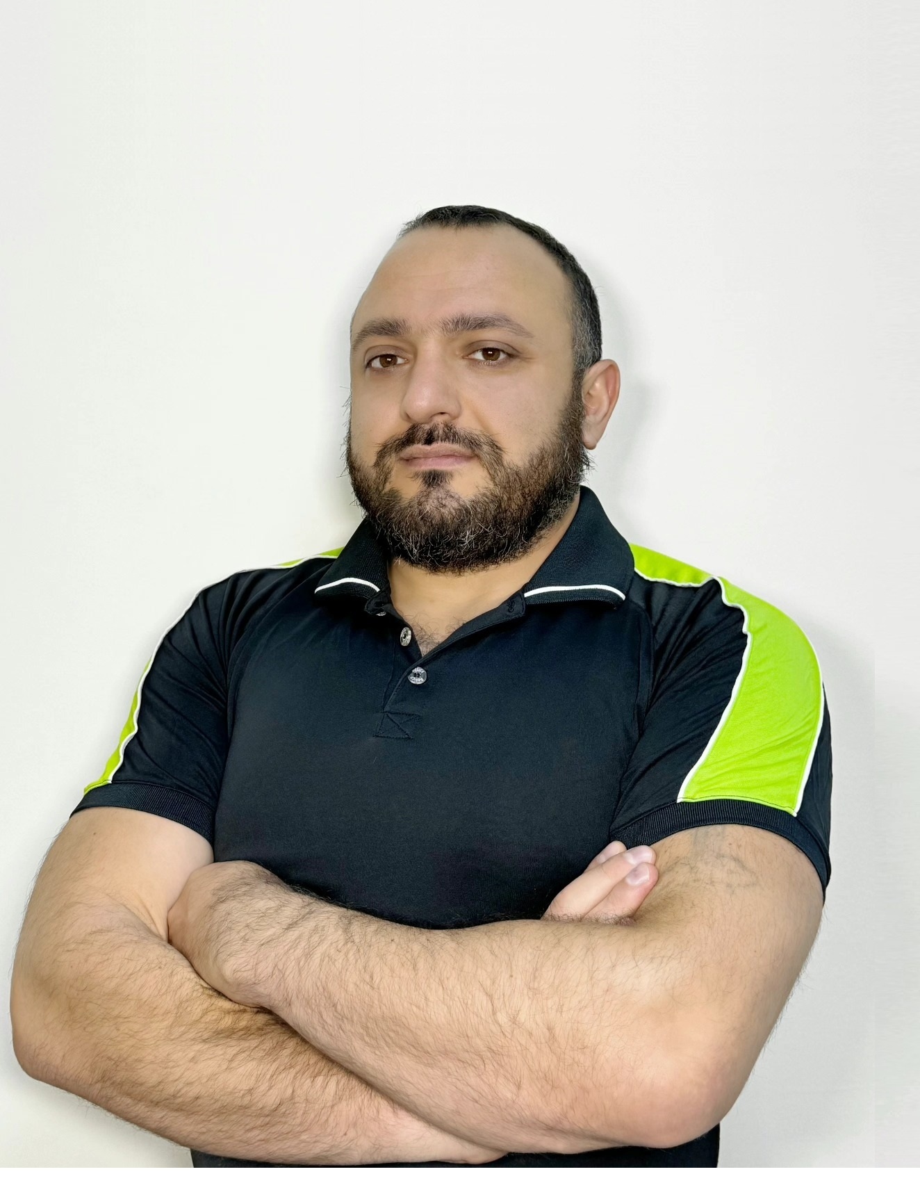 Portrait of Mushegh Manukyan wearing a dark polo shirt with bright green shoulder panels, standing with his arms crossed against a light background.